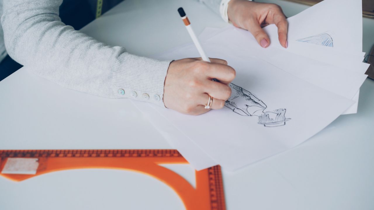 Vue rapprochée d’un artiste créant des croquis de mode, assis à un bureau en intérieur.